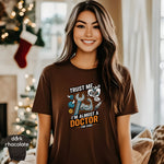 Trust Me I’m Almost a Doctor for Cars | Funny Mechanic T-Shirt worn by a smiling woman near a holiday backdrop.