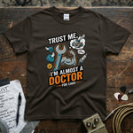 Trust Me I’m Almost a Doctor for Cars | Funny Mechanic T-Shirt displayed on a brown tee with wrench and mechanic graphics.