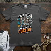 Trust Me I’m Almost a Doctor for Cars | Funny Mechanic T-Shirt in gray featuring a muscular arm and tools.